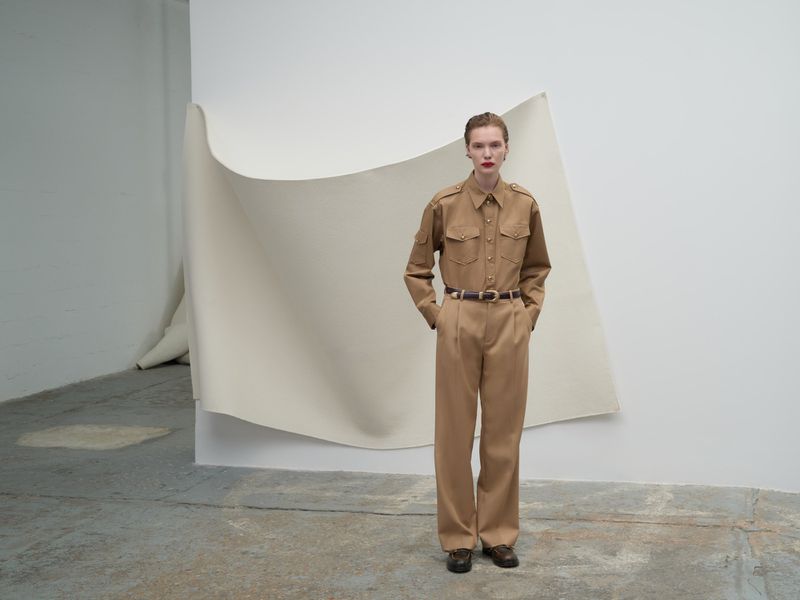 A fair-skinned woman in a tan jumpsuit and belt poses in a studio against a white backdrop in a medium shot with hands in pockets.