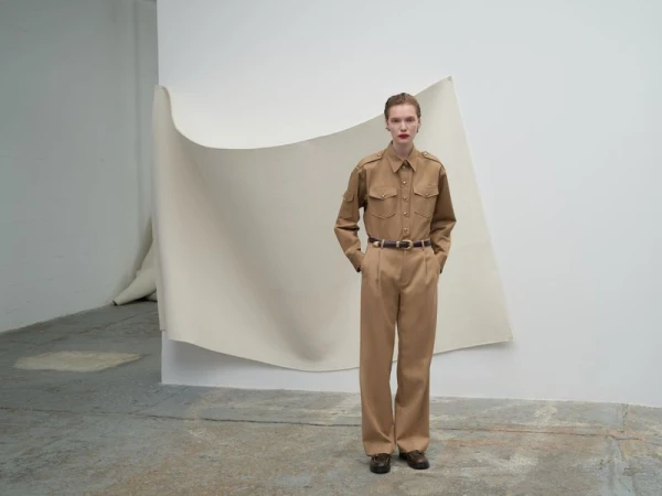 Model wearing a tan button-up shirt and matching trousers standing against a curved paper backdrop