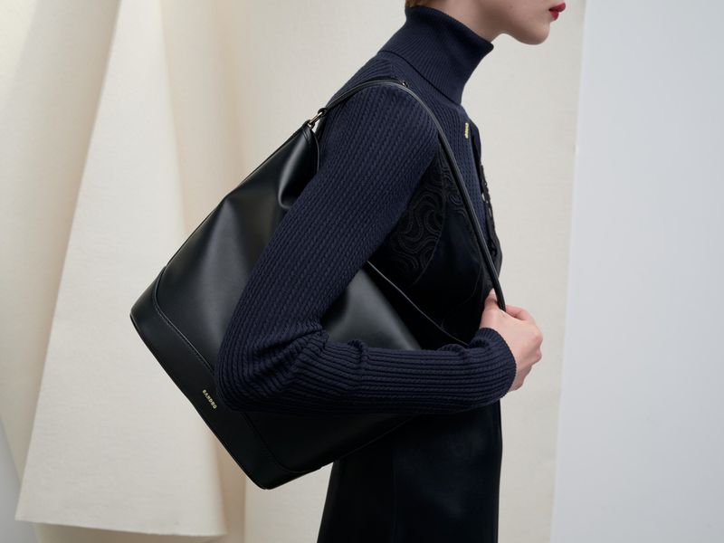 A woman with dark hair carries a large black leather shoulder bag in a studio setting with a minimalist background.