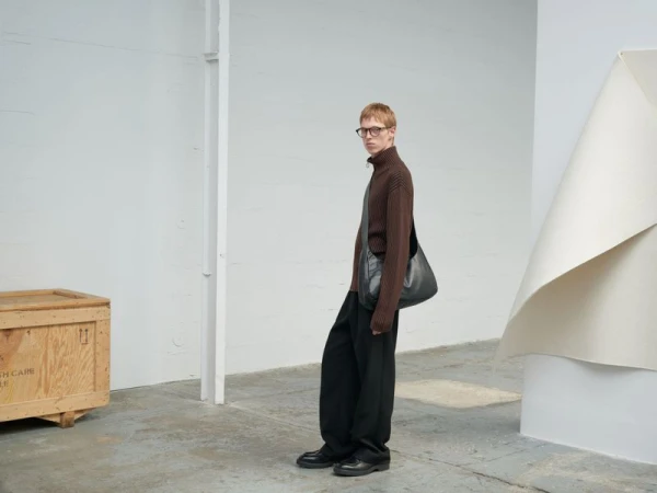 Male fashion model in a brown turtleneck and black pants poses with a leather messenger bag against a minimalist studio background.