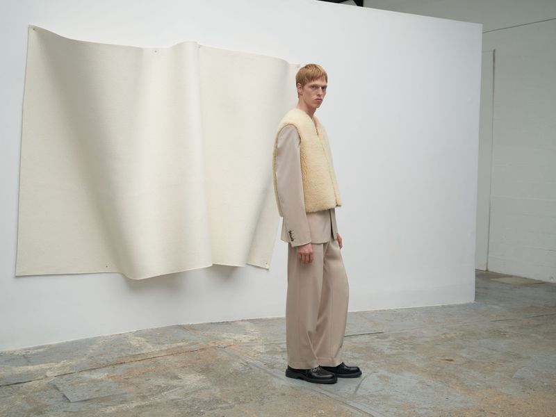 A fair-skinned man in a beige suit and sheepskin vest stands in a studio with a paper backdrop. Full shot, fashion editorial.