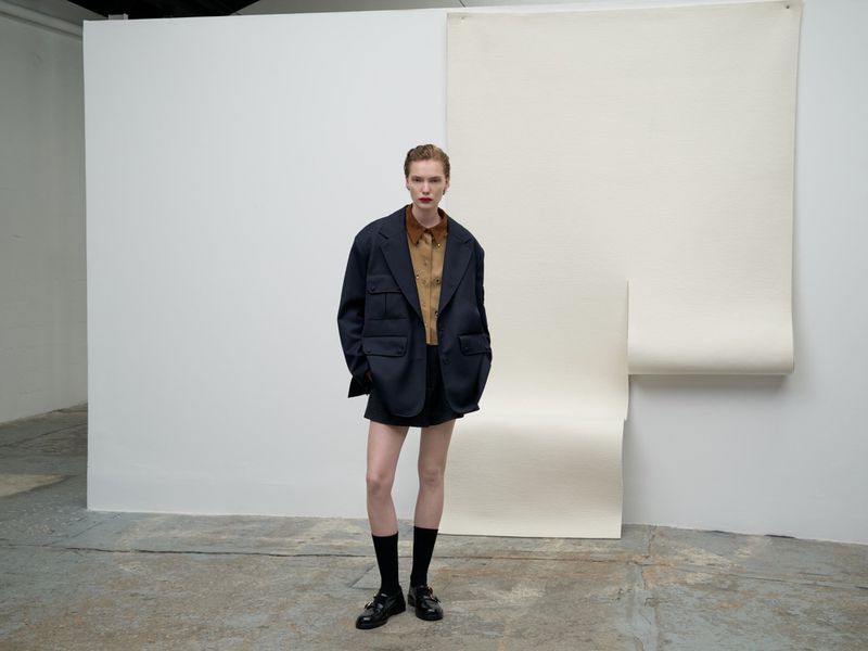 Full-length studio shot of an adult woman in an oversized blazer, brown shirt, and black shorts with buckled shoes against a white backdrop.