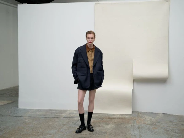 Full-length studio shot of an adult woman in an oversized blazer, brown shirt, and black shorts with buckled shoes against a white backdrop.