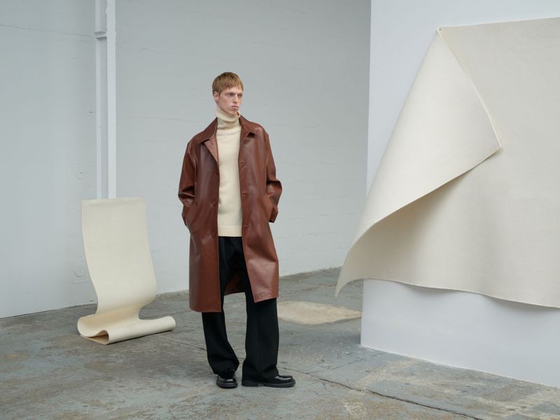 A fair-skinned man with short red hair poses in a brown leather overcoat, cream sweater, and black slacks in a minimalist studio.