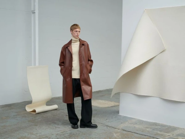 A fair-skinned man with short red hair poses in a brown leather overcoat, cream sweater, and black slacks in a minimalist studio.