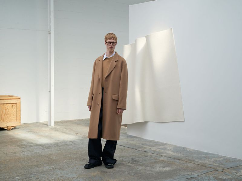 A blond male model wearing glasses, a tan coat and sweater, and black pants poses in a minimalist photo studio setting with a modern backdrop.