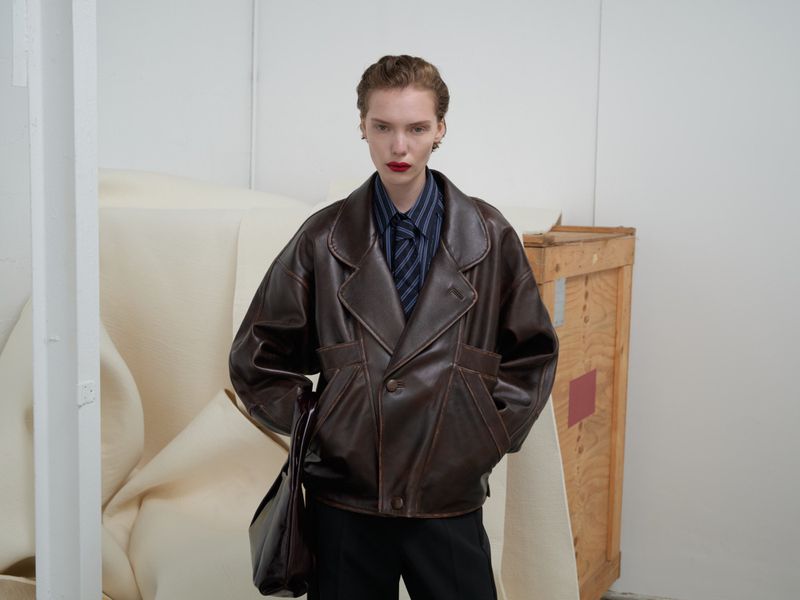 Stylish woman in a brown leather jacket, striped shirt, and dark pants poses in a photo studio with soft, neutral backdrop and wooden crate.