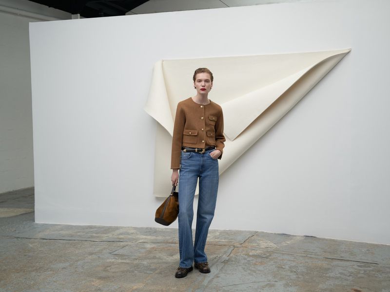 Fashionable woman in brown jacket and jeans stands in a bright studio near white abstract art, holding a brown leather purse.