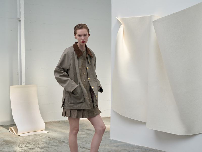 A slender woman with red lipstick models a brown jacket, matching pleated skirt, and button-down shirt in a minimalist indoor setting.