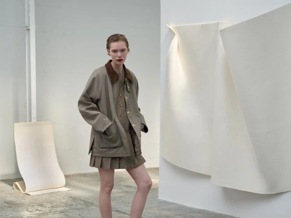 A slender woman with red lipstick models a brown jacket, matching pleated skirt, and button-down shirt in a minimalist indoor setting.