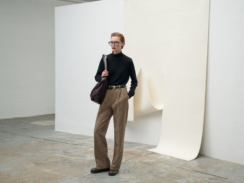 Adult woman poses in black turtleneck, brown trousers, and glasses against a neutral backdrop in a minimalist, editorial fashion shot.