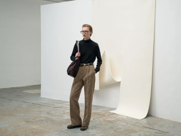 Adult woman poses in black turtleneck, brown trousers, and glasses against a neutral backdrop in a minimalist, editorial fashion shot.