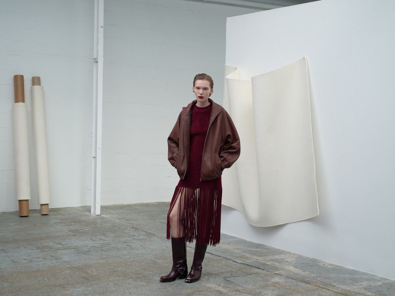 A fashionably dressed woman stands in a minimalist studio wearing a brown jacket, maroon dress with fringe, and knee-high boots.