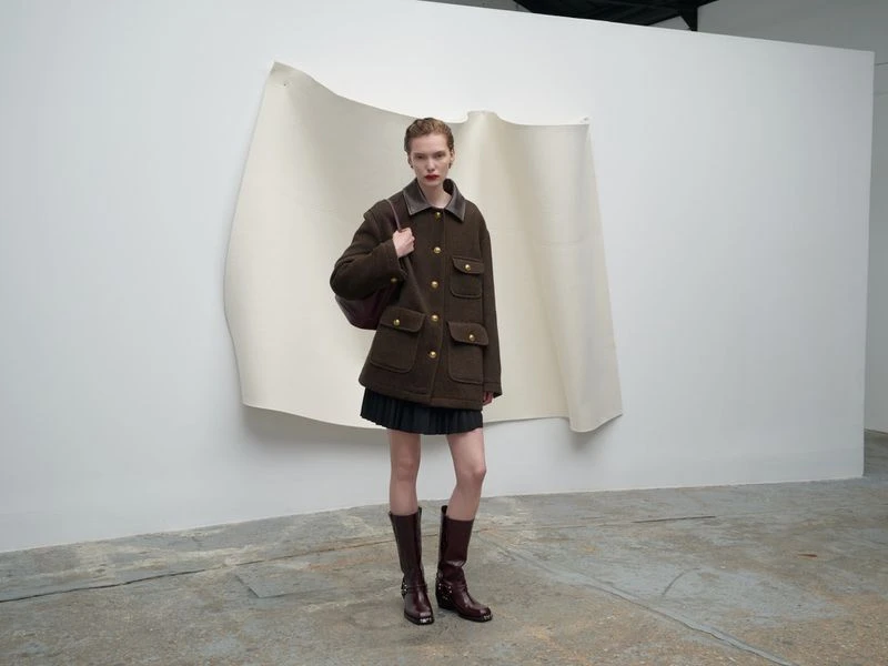 Full shot of a woman wearing an olive green jacket, dark skirt, and brown boots, posing in front of a white backdrop in a studio.