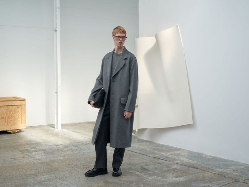 A fair-skinned male model in a grey coat and trousers stands in a photo studio holding a clutch bag, wearing glasses, in a neutral pose.