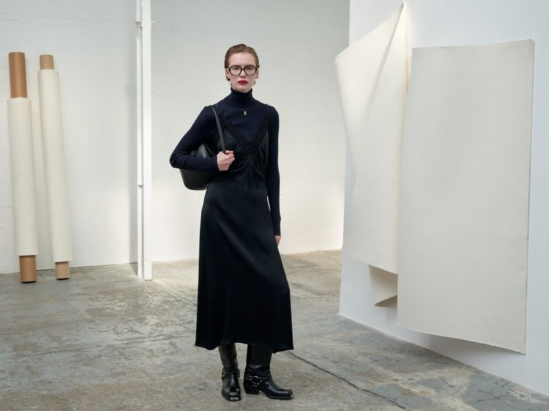 Full-length fashion shot of a woman in a black dress, turtleneck, boots, and glasses standing indoors against a white wall.