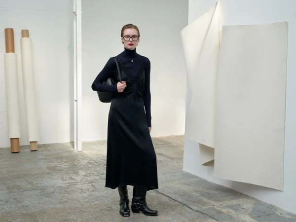 Full-length fashion shot of a woman in a black dress, turtleneck, boots, and glasses standing indoors against a white wall.