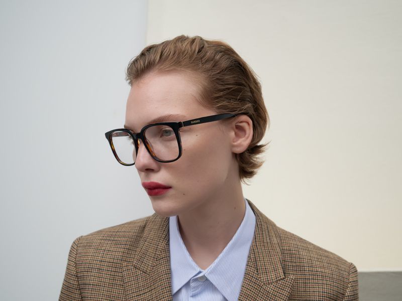 Close-up of a brunette adult woman wearing a blazer and large black-rimmed glasses, posing in front of a plain white wall.