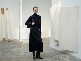 Model in a black slip dress over a dark turtleneck holding a handbag in a minimalist white studio space