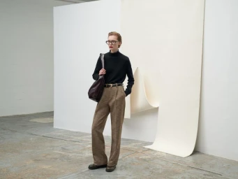 Model wearing black turtleneck and brown patterned trousers holding a dark handbag against a white backdrop.