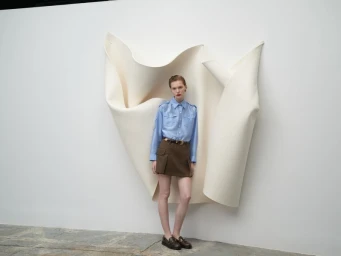 Model wearing a light blue button-down shirt and brown miniskirt standing against a white wall with large sculptural paper art backdrop