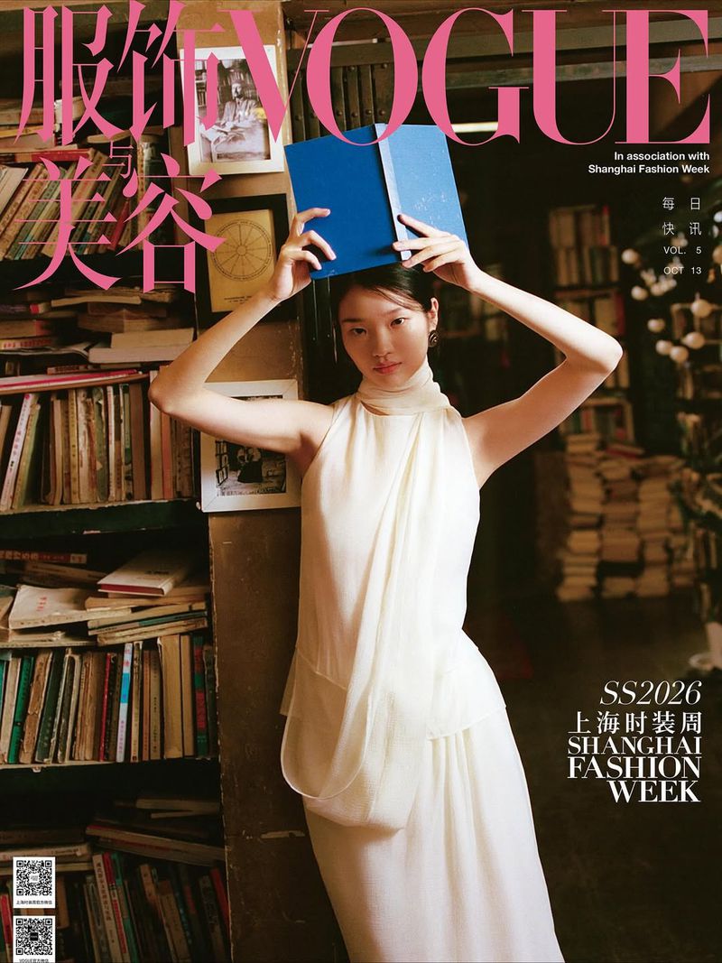 Vogue cover featuring a woman in white dress holding a blue book over her head, posing in front of a shelf full of books.