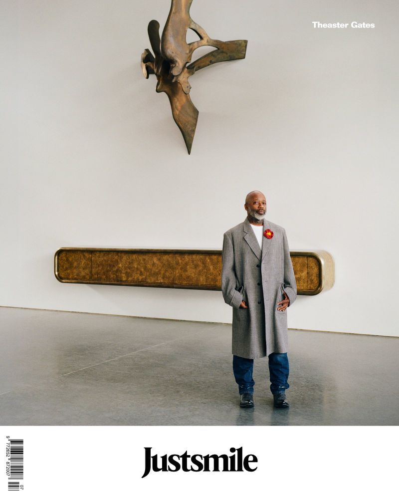 Adult male in a grey coat stands in an art gallery with a metal sculpture and a gold wall installation against a white wall and grey floor.