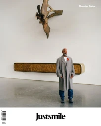 Theaster Gates standing in a minimalist gallery wearing a grey coat and blue jeans beneath a wall sculpture.