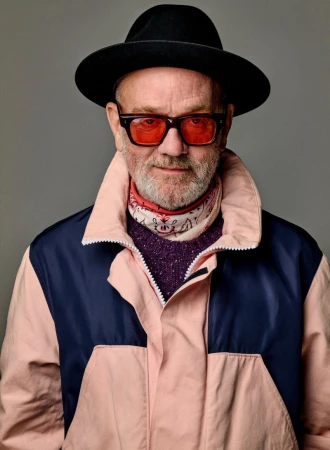 Stylish adult male portrait wearing a hat, orange sunglasses, and a pink jacket against a grey background in a photo studio.