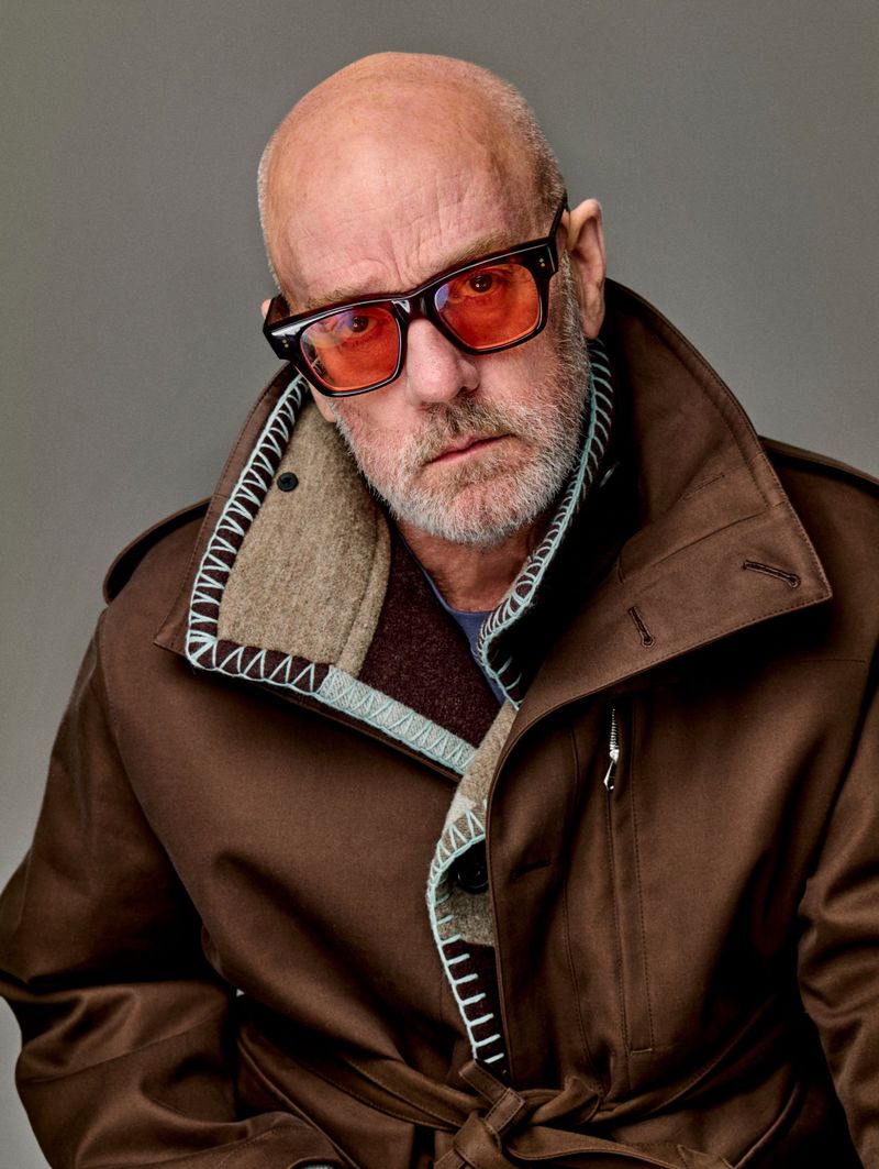Portrait of a mature bald man wearing glasses and a brown coat in a studio setting, looking directly at the camera with a neutral expression.
