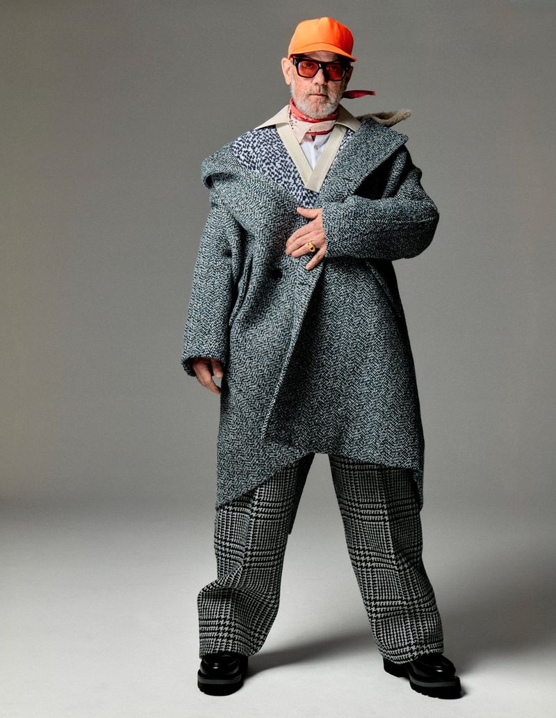 Full-length shot of a mature adult male wearing an oversized gray coat, checkered pants, a red neck scarf, and an orange baseball cap in studio.
