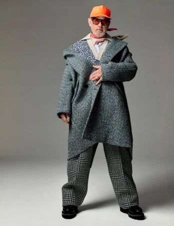 Full-length shot of a mature adult male wearing an oversized gray coat, checkered pants, a red neck scarf, and an orange baseball cap in studio.