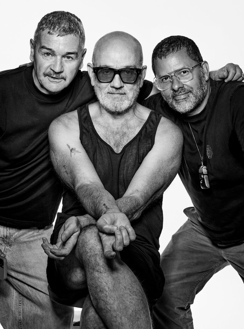 Three adult males pose in a photo studio. The image is black and white, featuring varied facial hair, dark tank top, and light-colored pants.
