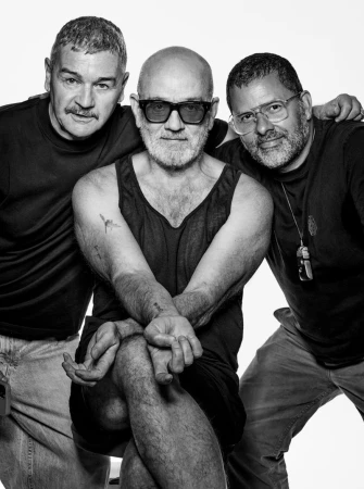 Three adult males pose in a photo studio. The image is black and white, featuring varied facial hair, dark tank top, and light-colored pants.