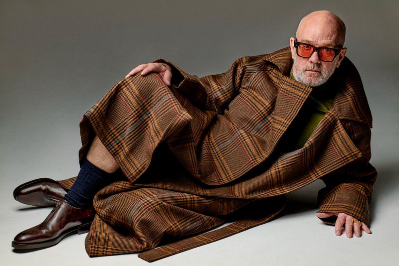 Portrait of a bald adult man in a brown plaid coat, orange sunglasses, and brown shoes, lying on a grey surface in a studio.
