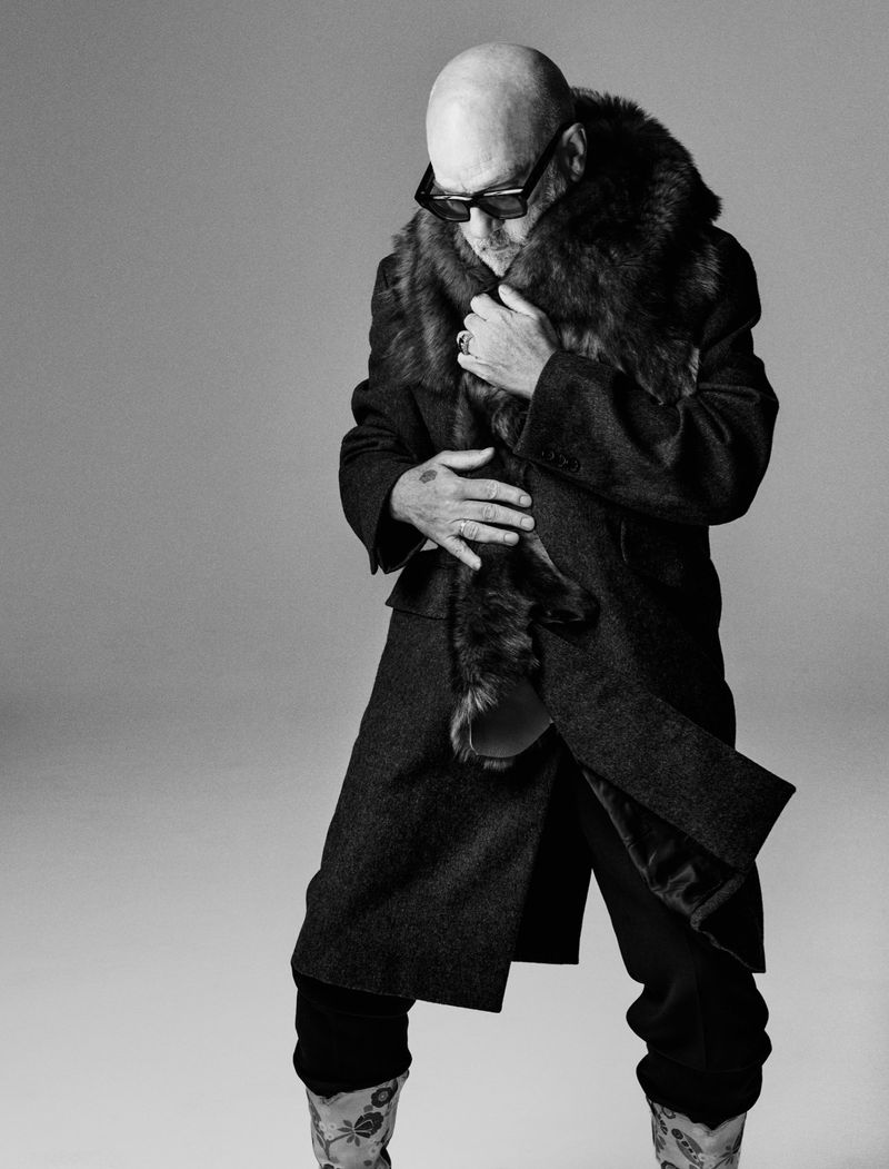 Black and white portrait of a bald adult male wearing a coat with a fur collar and patterned boots posing in a photo studio.