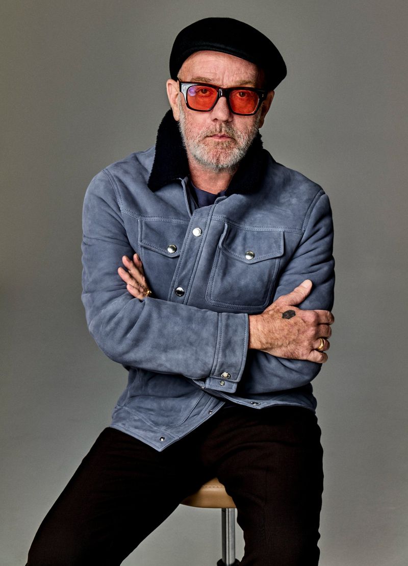 Portrait of a mature adult male wearing red eyeglasses, a blue jacket with a dark collar, and a black cap, posed indoors on a stool.