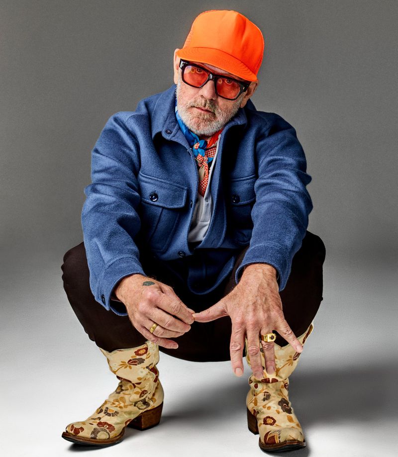 Stylish adult male in blue jacket and floral boots squats in a studio, wearing an orange baseball cap and red glasses. Neutral backdrop.
