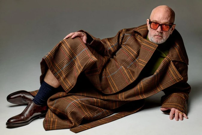 Michael Stipe reclining on a grey studio backdrop wearing a plaid brown coat, tinted glasses, and dark dress shoes.