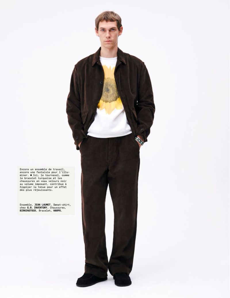 A slender adult male poses in a full-length shot, wearing a brown corduroy jacket and pants with a white graphic tee in a studio.