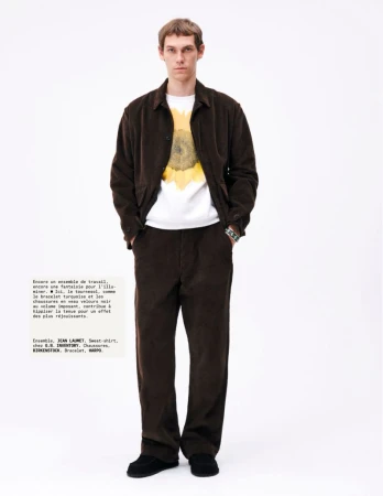 A slender adult male poses in a full-length shot, wearing a brown corduroy jacket and pants with a white graphic tee in a studio.
