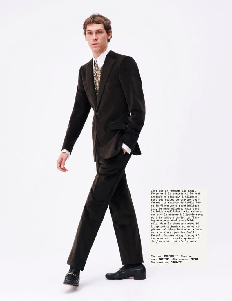Adult male model wearing a dark brown corduroy suit, white shirt, and patterned tie walks on a white background in a studio shot.