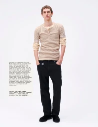 Full-length portrait of a male model wearing layered beige henleys and black trousers against a white seamless backdrop.