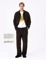 Full-length portrait of a male model wearing a dark brown corduroy jacket and matching trousers over a white t-shirt with a sunflower graphic.