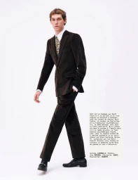 Full-length portrait of a male model wearing a dark corduroy suit, patterned tie, and loafers against a plain white background.
