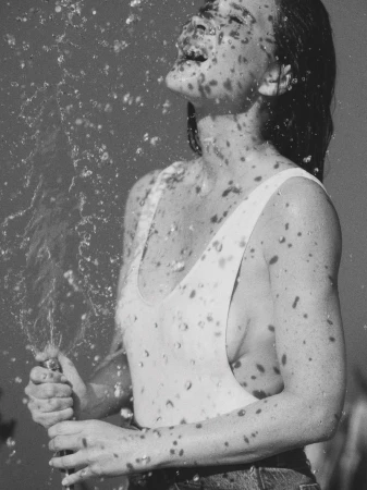 Monochrome shot of a woman in a swimsuit being splashed with water, looking up with her mouth open in an outdoor environment.