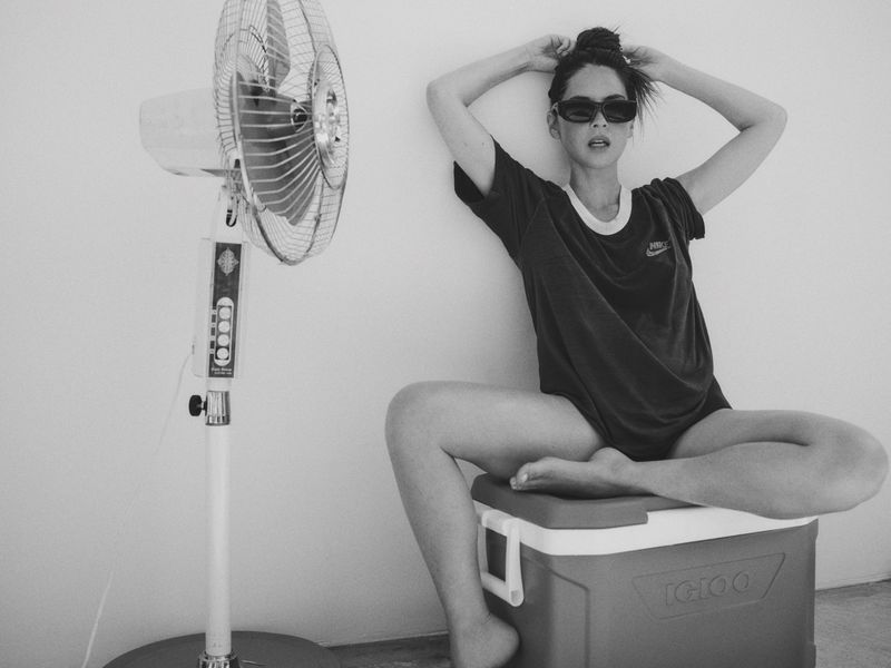 Black and white portrait of a woman sitting on a cooler with a fan in the background, posing with hands on her head and sunglasses.
