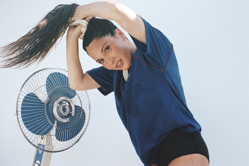 Brunette woman in a blue shirt and black shorts is holding up her hair in front of an electric fan against a pale blue background.