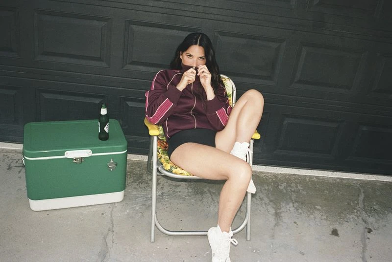 Woman sitting on a yellow-and-white lawn chair with a beer cooler and beer bottle next to her in front of a garage door.