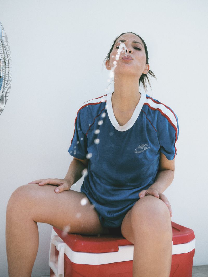 Woman in blue sports jersey seated on a cooler, blowing water from her mouth in front of a fan indoors with a white background.